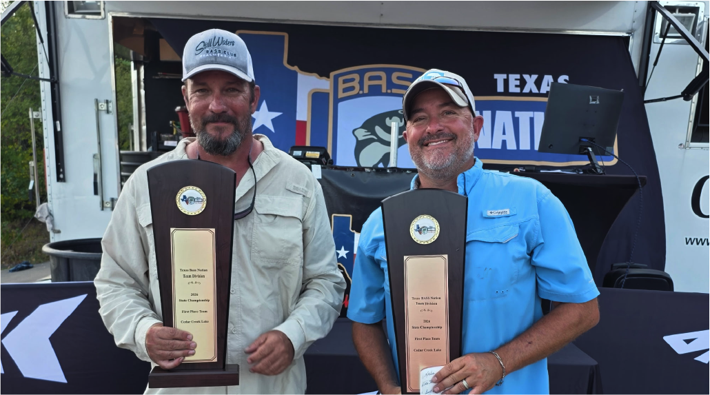 Two men standing outdoors in front of a truck and backdrop, each holding a tall trophy plaque. The man on the left wears a light beige shirt and a white cap, and the man on the right wears a light blue shirt and a white cap.