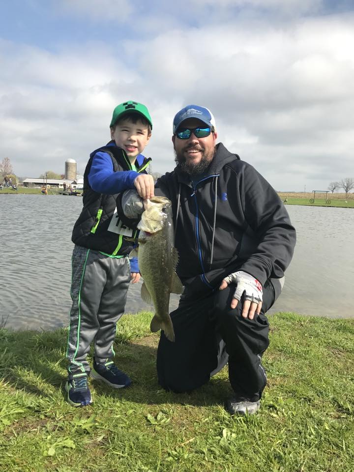 A man and a boy kneeling on grass by a lake, holding a large fish. The man wears a black jacket, black pants, sunglasses, and a blue cap, while the boy wears a green cap, a blue and black vest, and gray pants.