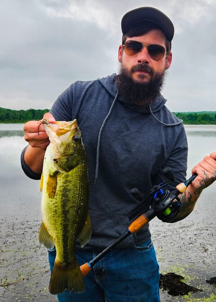 A bearded man wearing a black cap and sunglasses stands on a muddy lakeshore holding a freshly caught largemouth bass while holding a fishing rod. The background shows a calm lake and a green forest under an overcast sky.